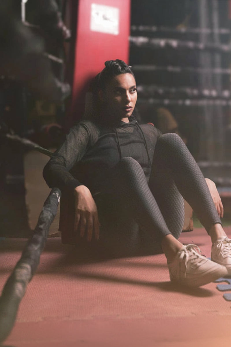 A woman sits on a gym floor wearing textured black leggings and white sneakers, with a red boxing ring visible in the background. The word 'LEGGINGS' appears in white text at the bottom left.